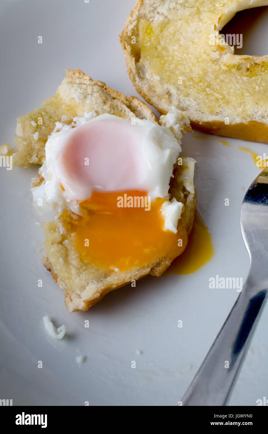 A partly eaten free range fried egg with yolk spilling onto bagel and plate below.  Fork visible.  Shot from diner's perspective. Stock Photo