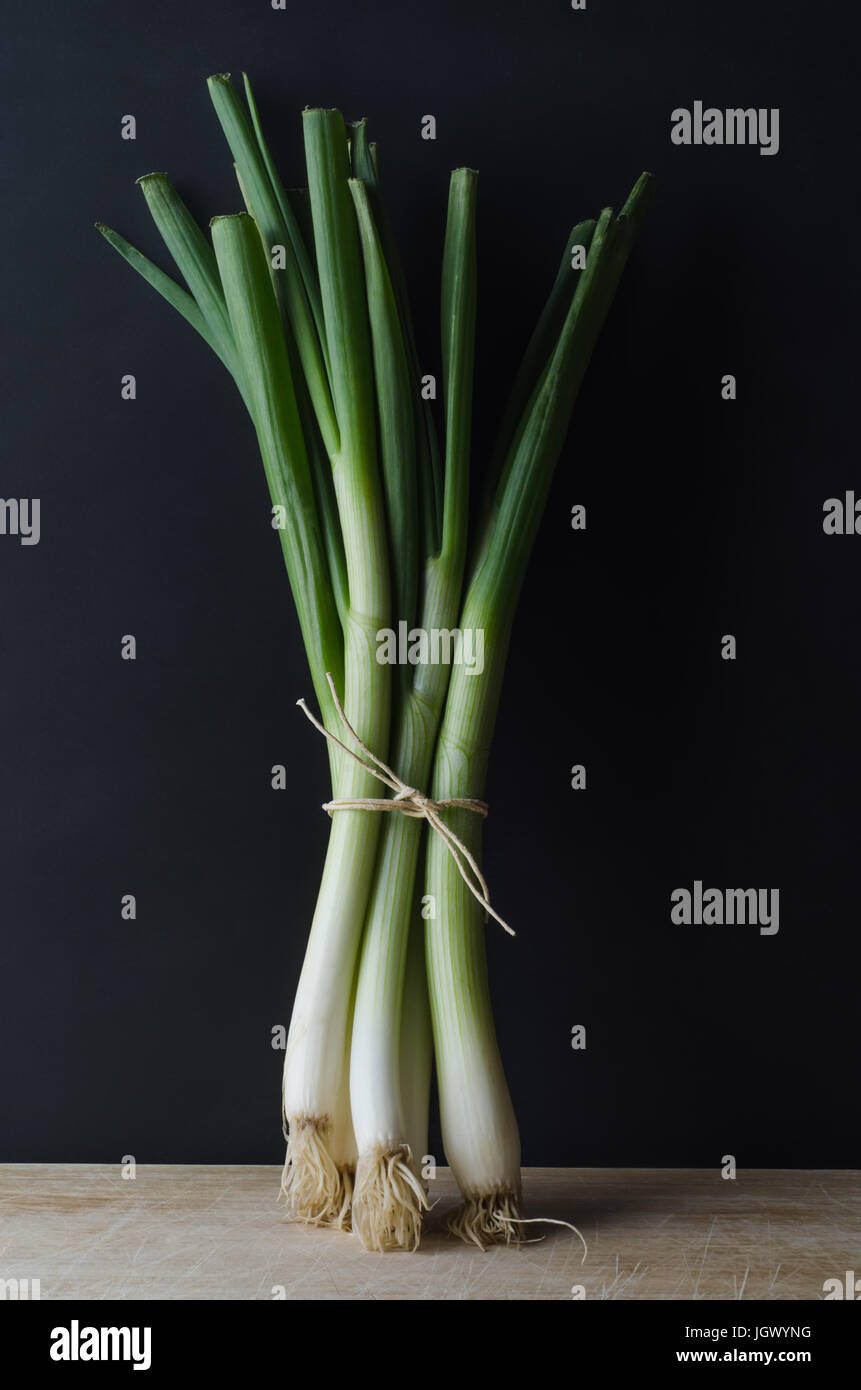 A bunch of spring onions (scallions) tied with string and standing upright on a scratched and worn wooden surface against a black chalkboard backgroun Stock Photo