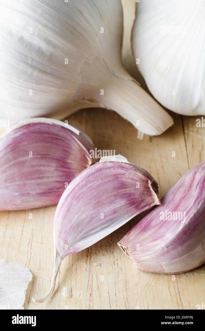 Elevated, close up view of whole, unpeeled garlic bulbs and three pink cloves, with scattered papery skin on a scratched wooden chopping  board. Stock Photo