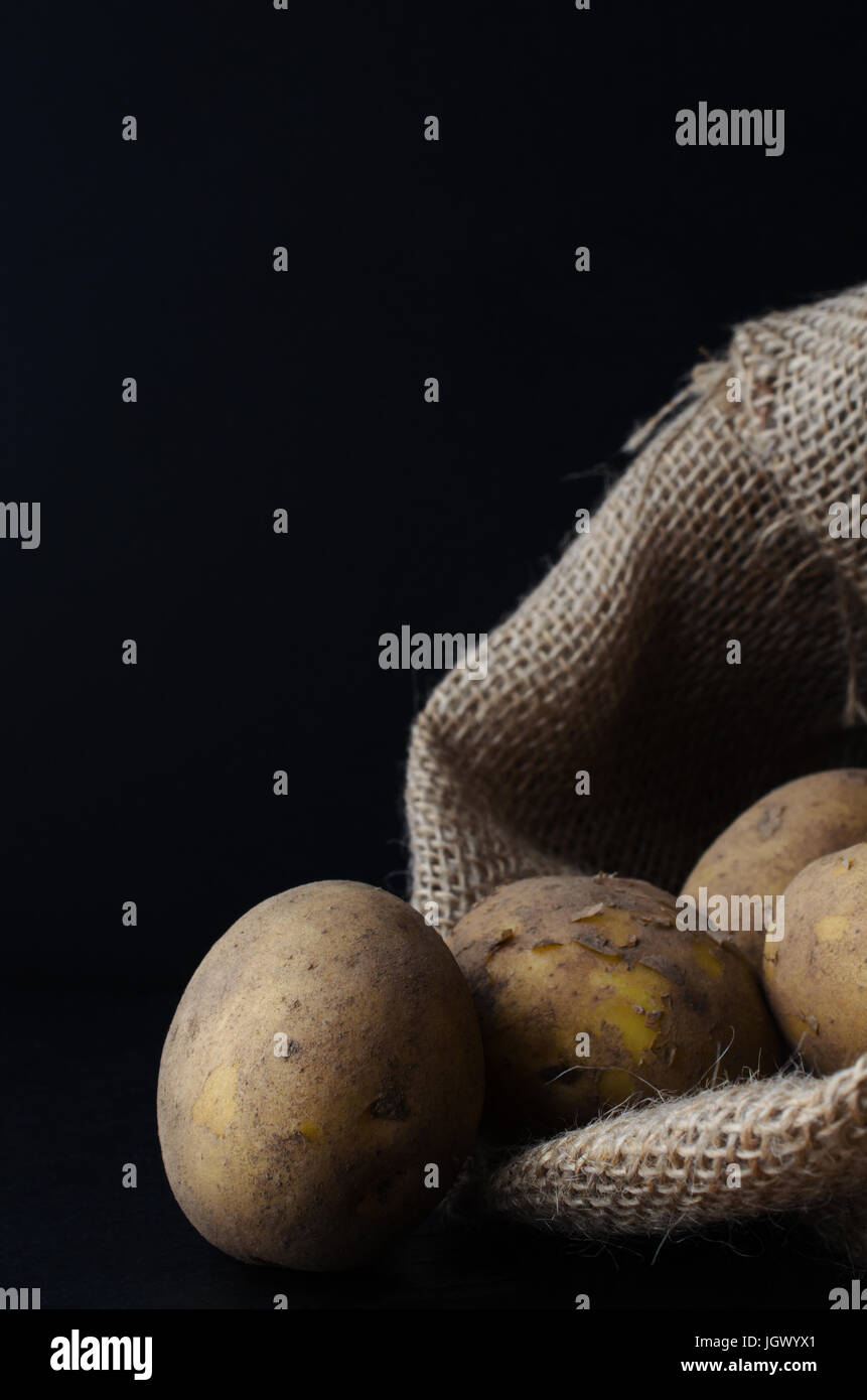 Raw, unwashed, unpeeled potatoes, spilling out of hessian sack.  Moody lighting, black surface and background. Stock Photo