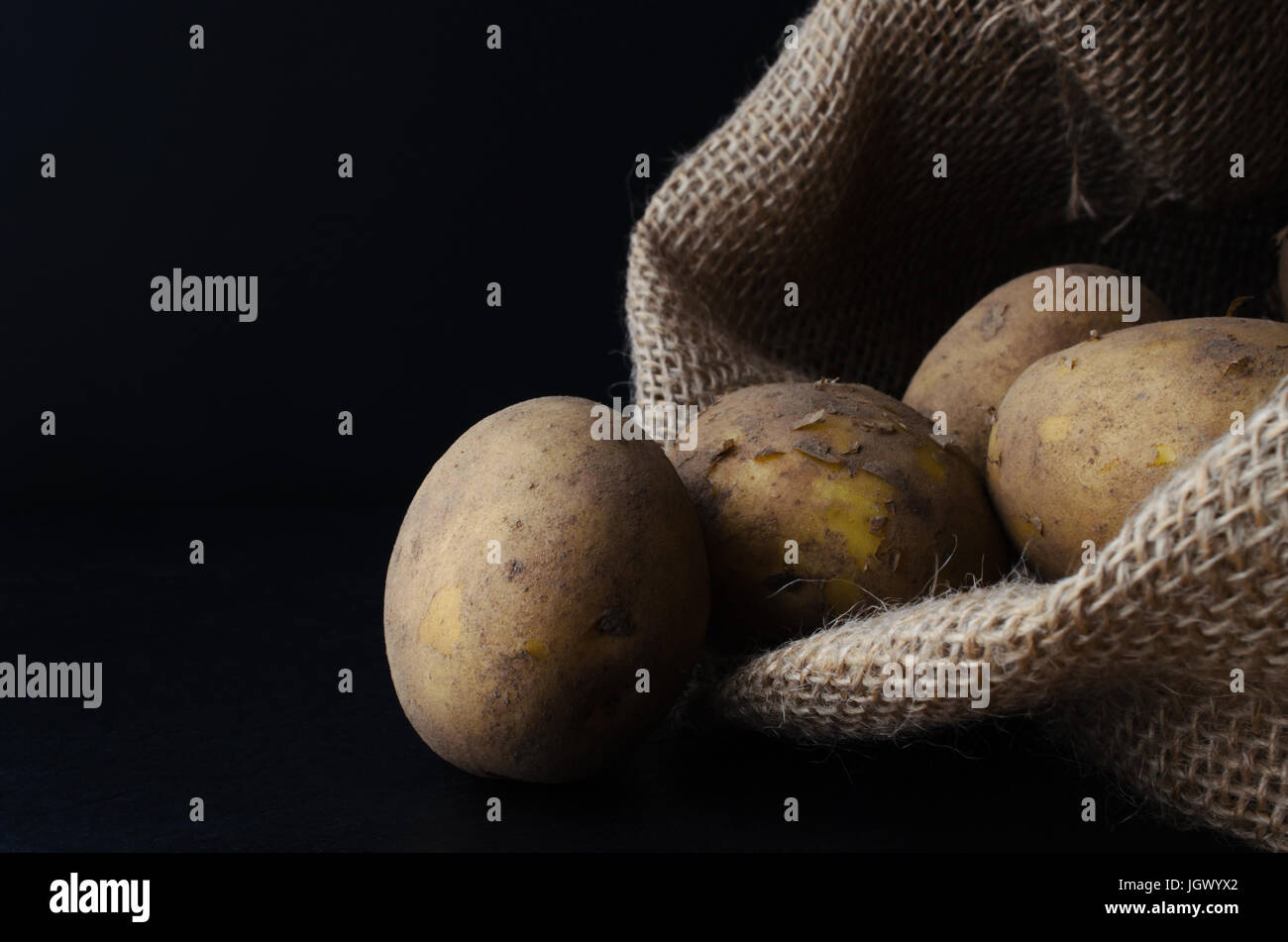 Raw, unwashed, unpeeled potatoes, spilling out of hessian sack on to black slate surface.  Moody lighting, black background. Stock Photo