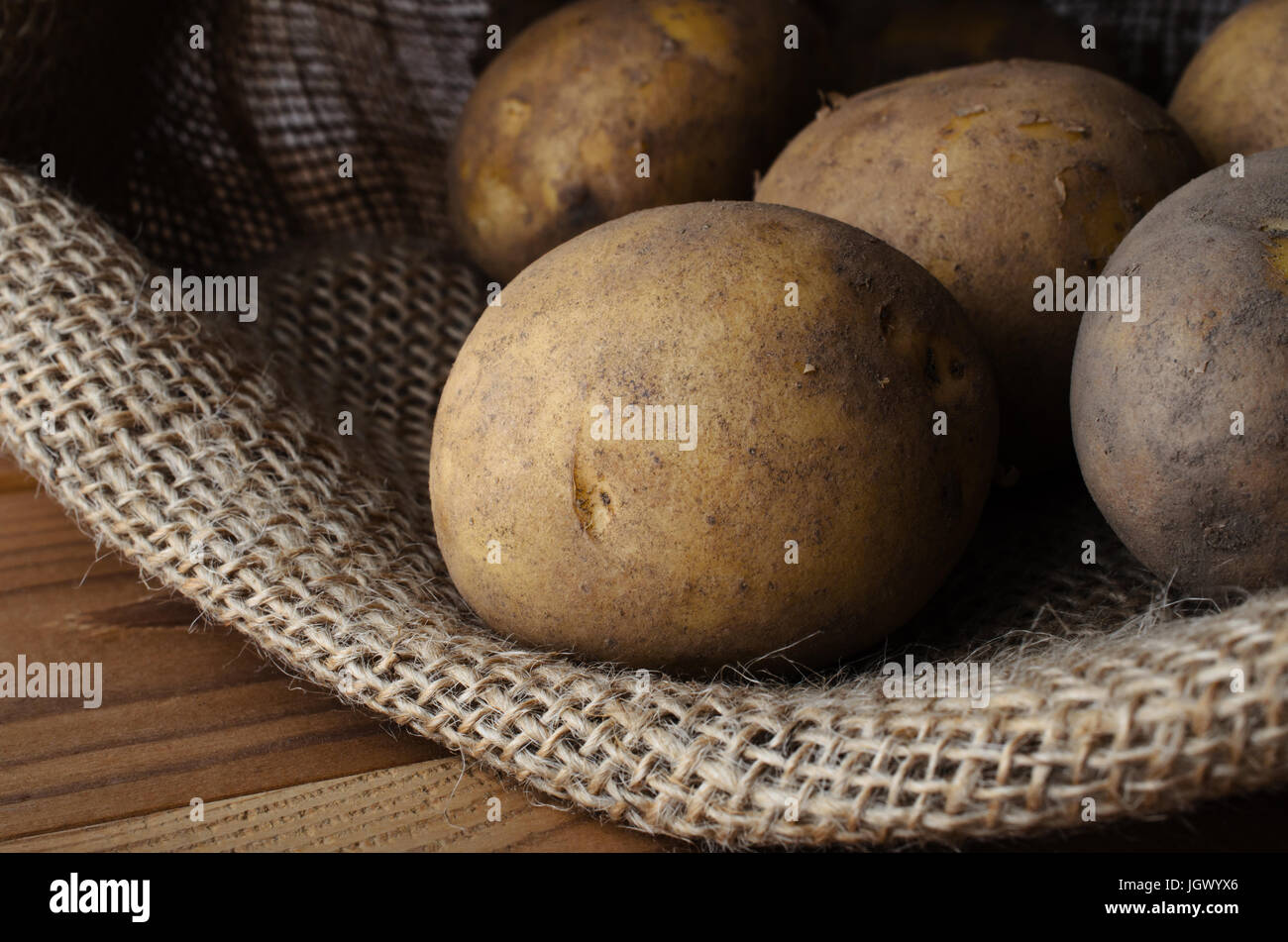 Raw, unwashed, unpeeled potatoes in opened hessian sack, lain on wood planked table. Stock Photo