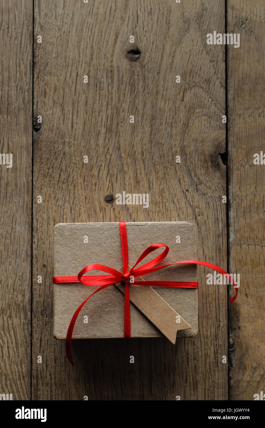 Overhead shot of a simple brown gift box on an old oak wood planked table, tied to a bow with red satin ribbon, with a blank vintage style parcel tag  Stock Photo
