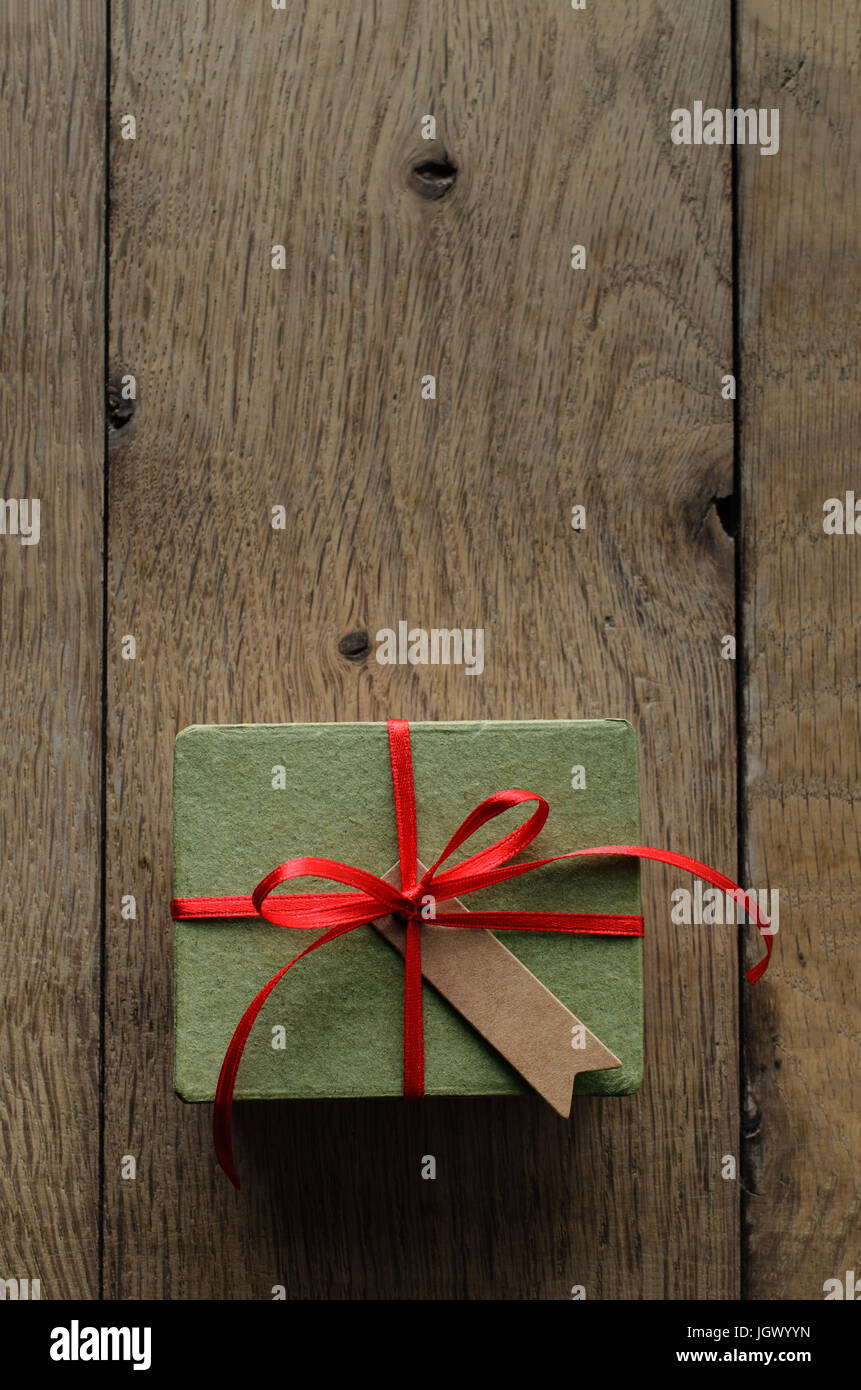 Overhead shot of a simple green gift box on an old oak wood planked table, tied to a bow with red satin ribbon, with a blank vintage style parcel tag  Stock Photo