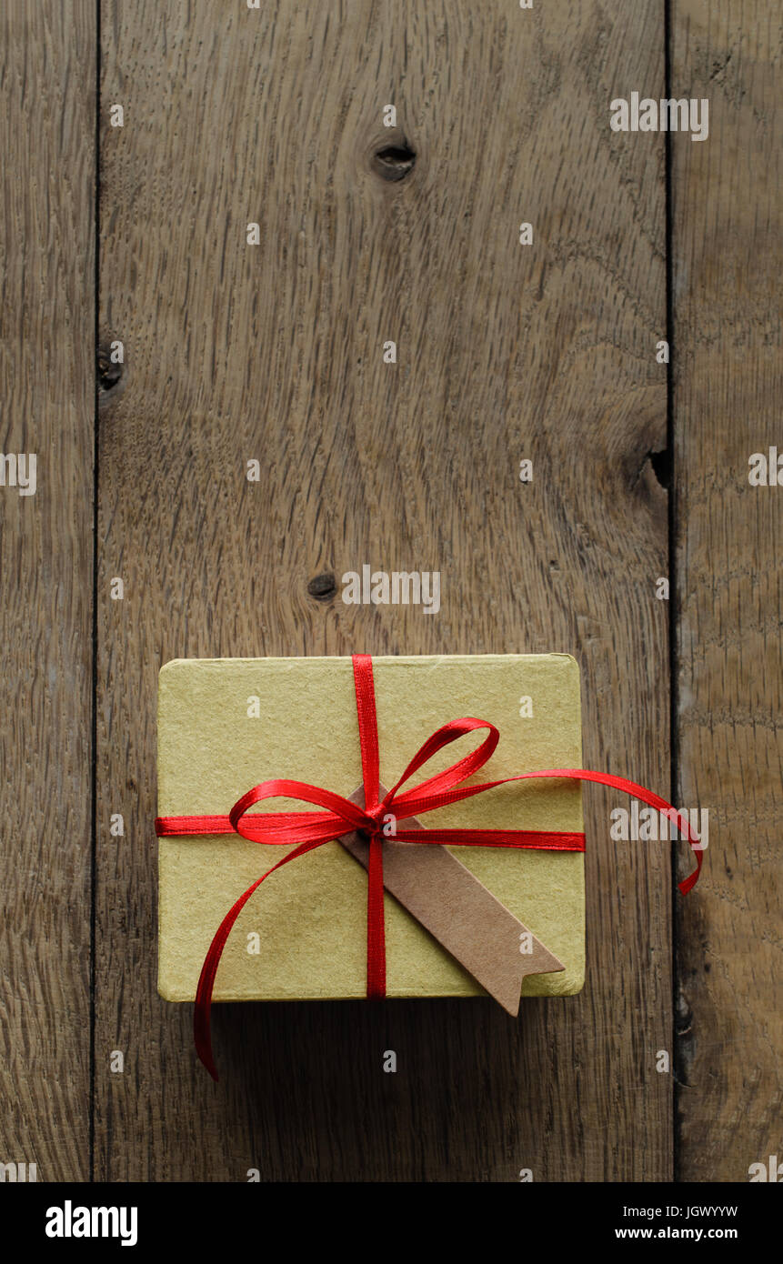 Overhead shot of a simple yellow gift box on an old oak wood planked table, tied to a bow with red satin ribbon, with a blank vintage style parcel tag Stock Photo