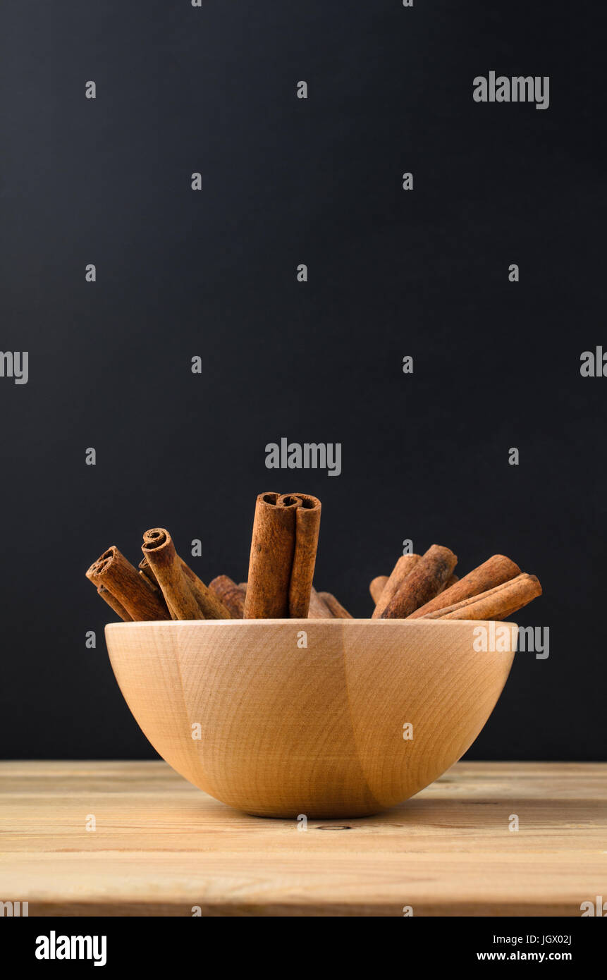 Culinary spice image of Cinnamon sticks, grouped in a wooden bowl on a light wood plank table with black chalkboard in background. Stock Photo