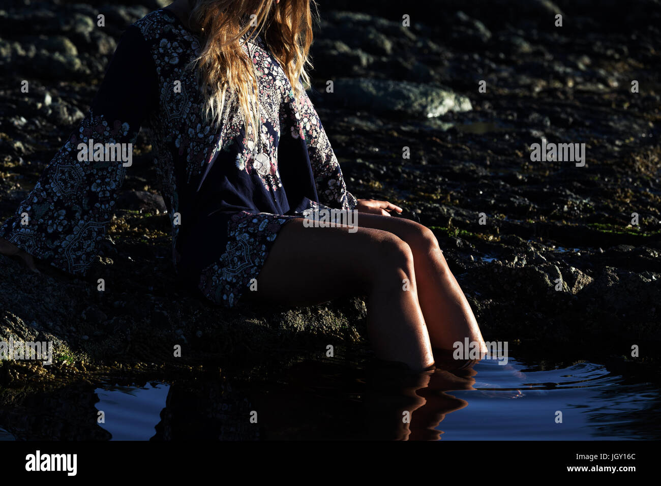 Stylish girl in dress sits by a rock pool at the beach Stock Photo