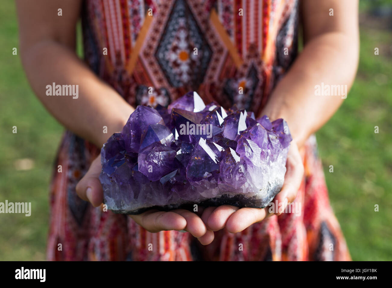A young woman holds a large cluster of amethyst crystals as they shimmer in the sunlight. Stock Photo