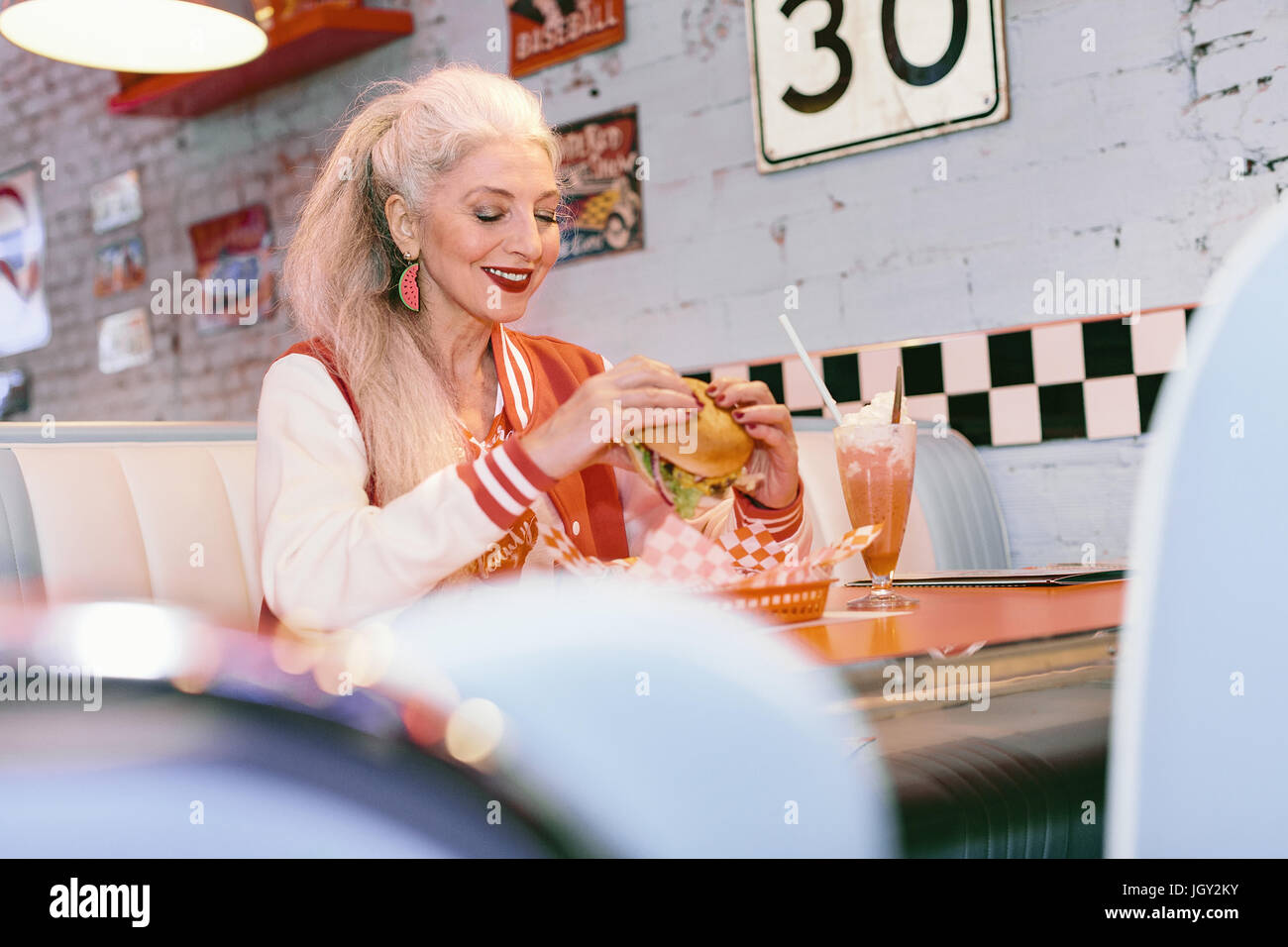 Mature woman in baseball jacket eating burger in 1950's diner Stock Photo