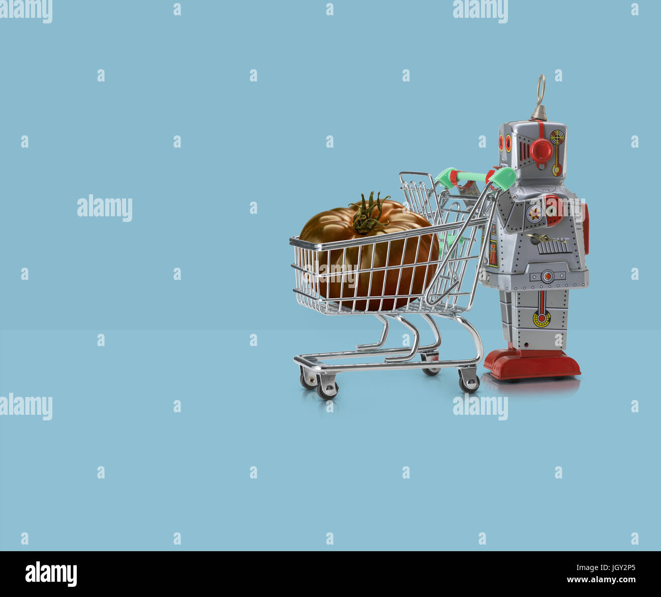 Toy robot pushing miniature shopping trolley with tomato against blue