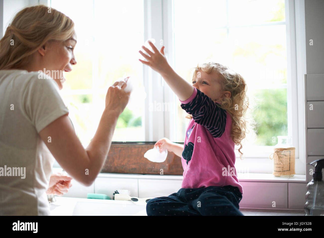 Mother and daughter in kitchen, daughter sitting on work surface playing with washing up bubbles Stock Photo