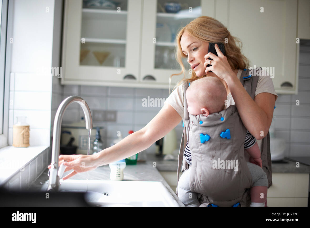 Woman holding baby boy in sling, using smartphone, turning on tap to do washing up Stock Photo