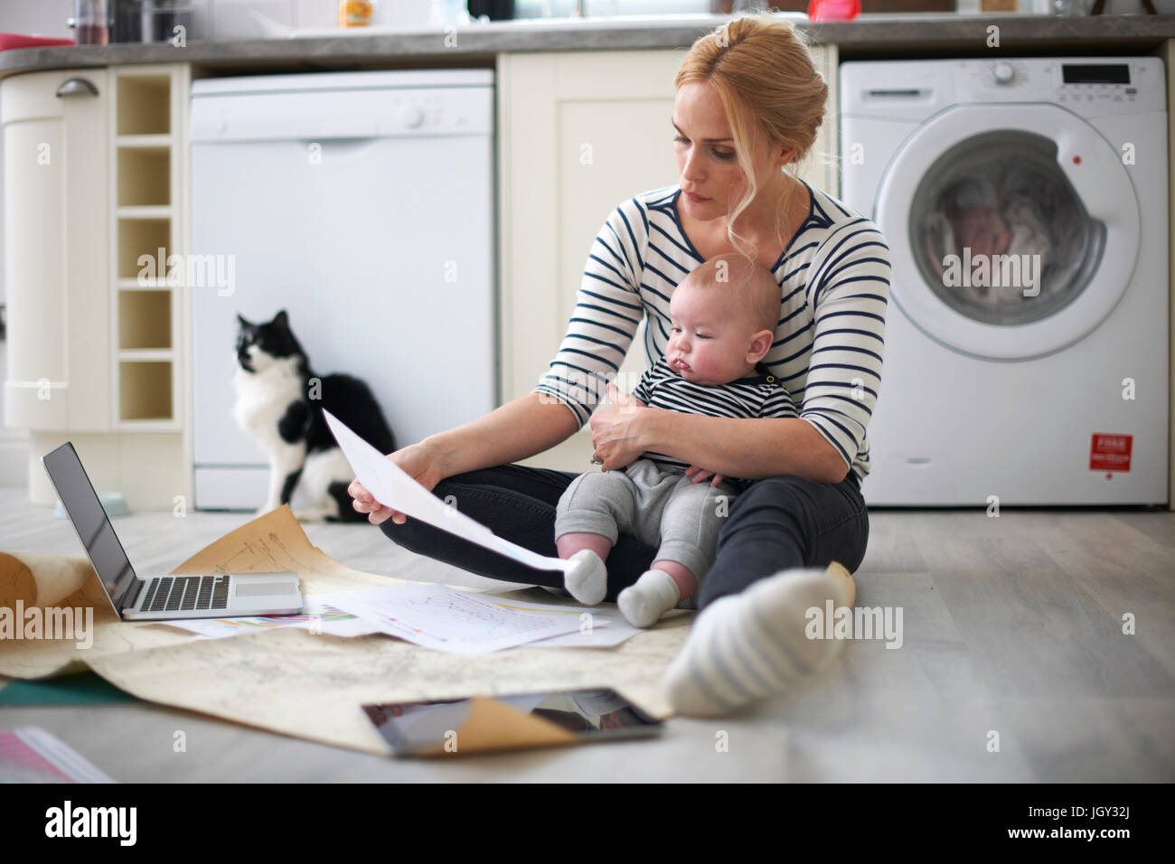 Woman holding baby son in arms, looking through work on kitchen floor, laptop and digital tablet in front of her Stock Photo