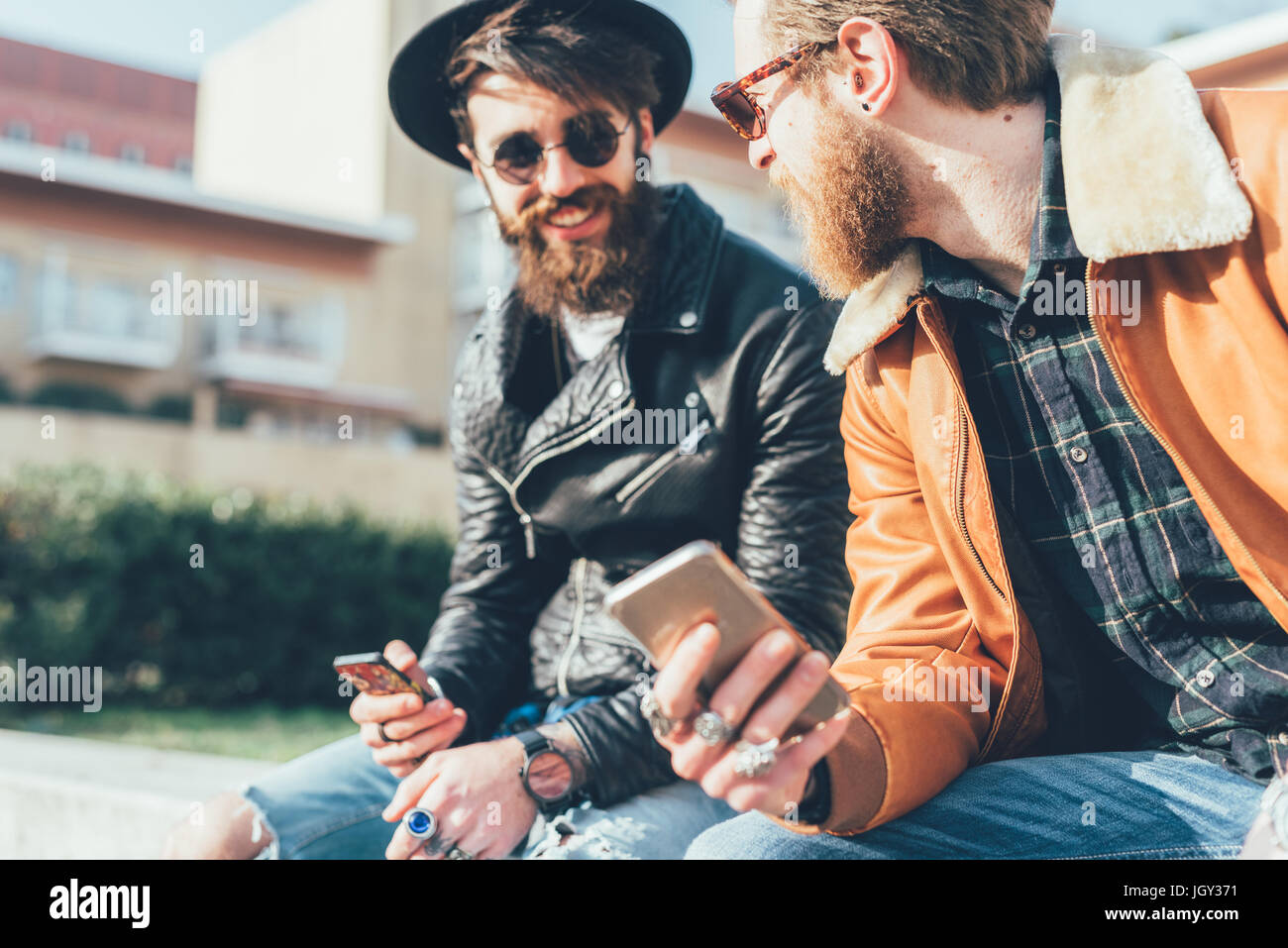 Two young male hipsters sitting on wall with smartphones Stock Photo