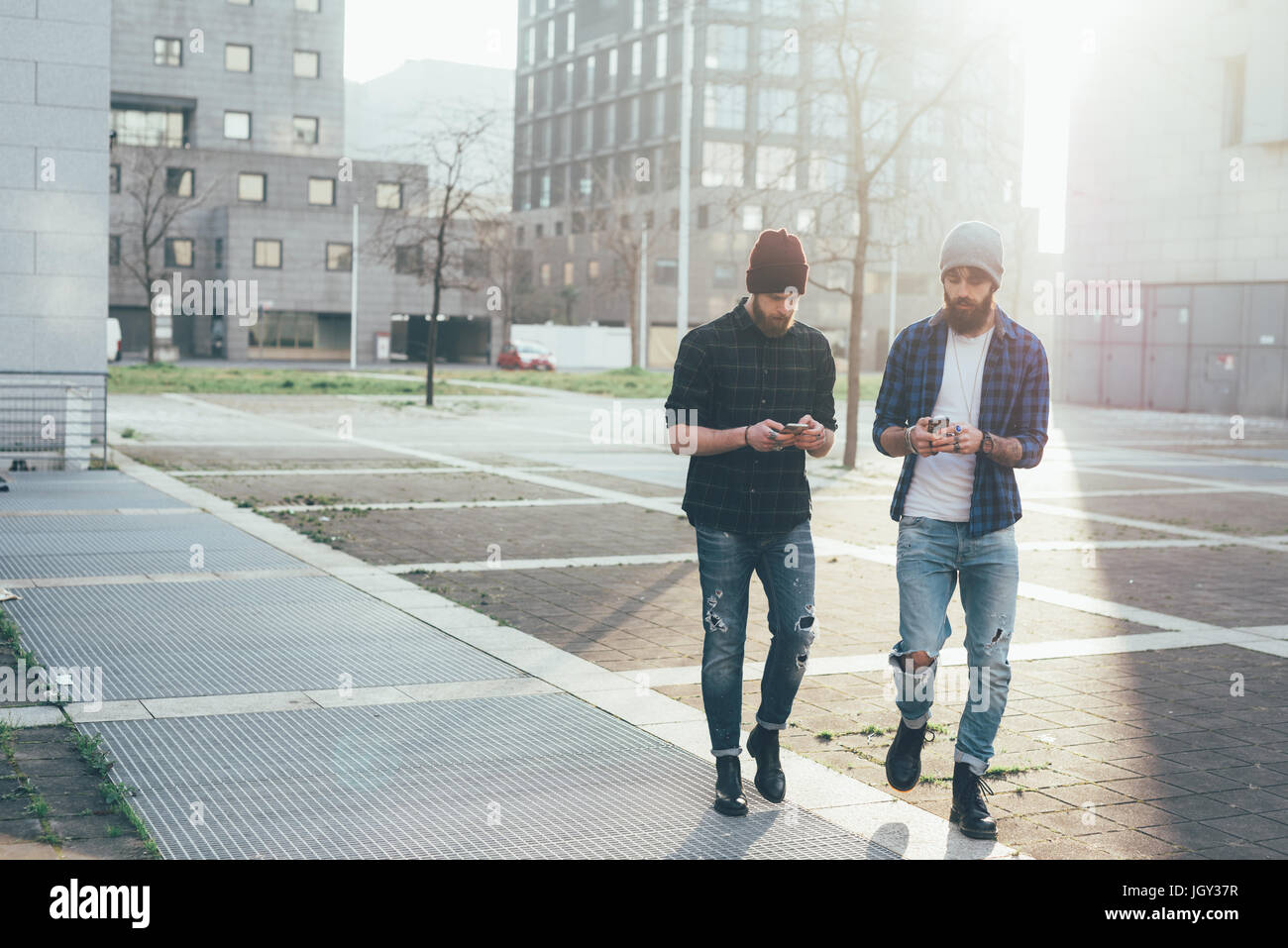 Two young male hipsters walking in city while looking at smartphones Stock Photo