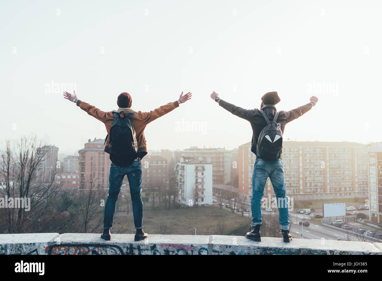Rear view of two young male hipsters standing on city wall with arms out Stock Photo