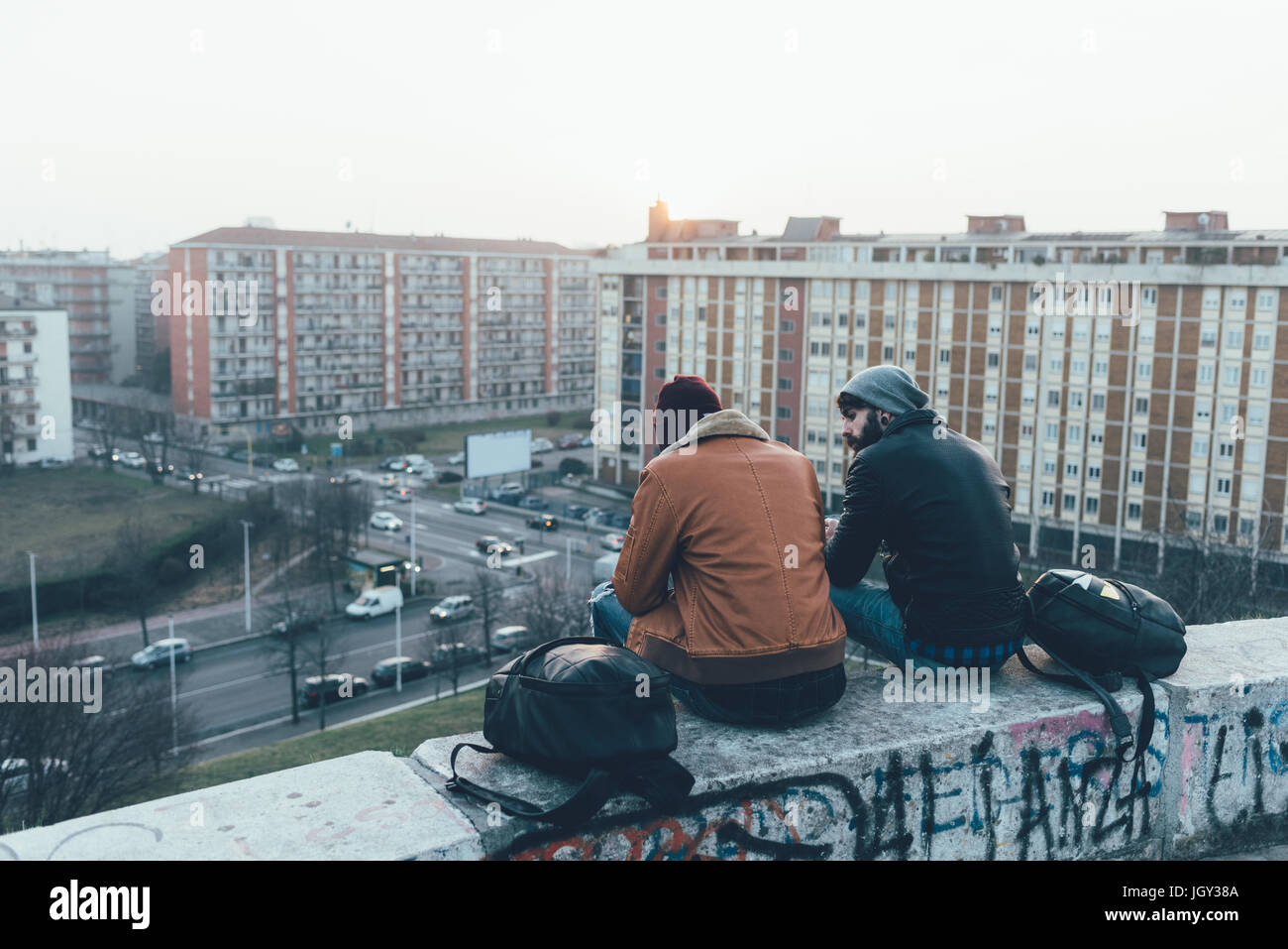 Rear view of two young male hipsters sitting on wall above cityscape Stock Photo