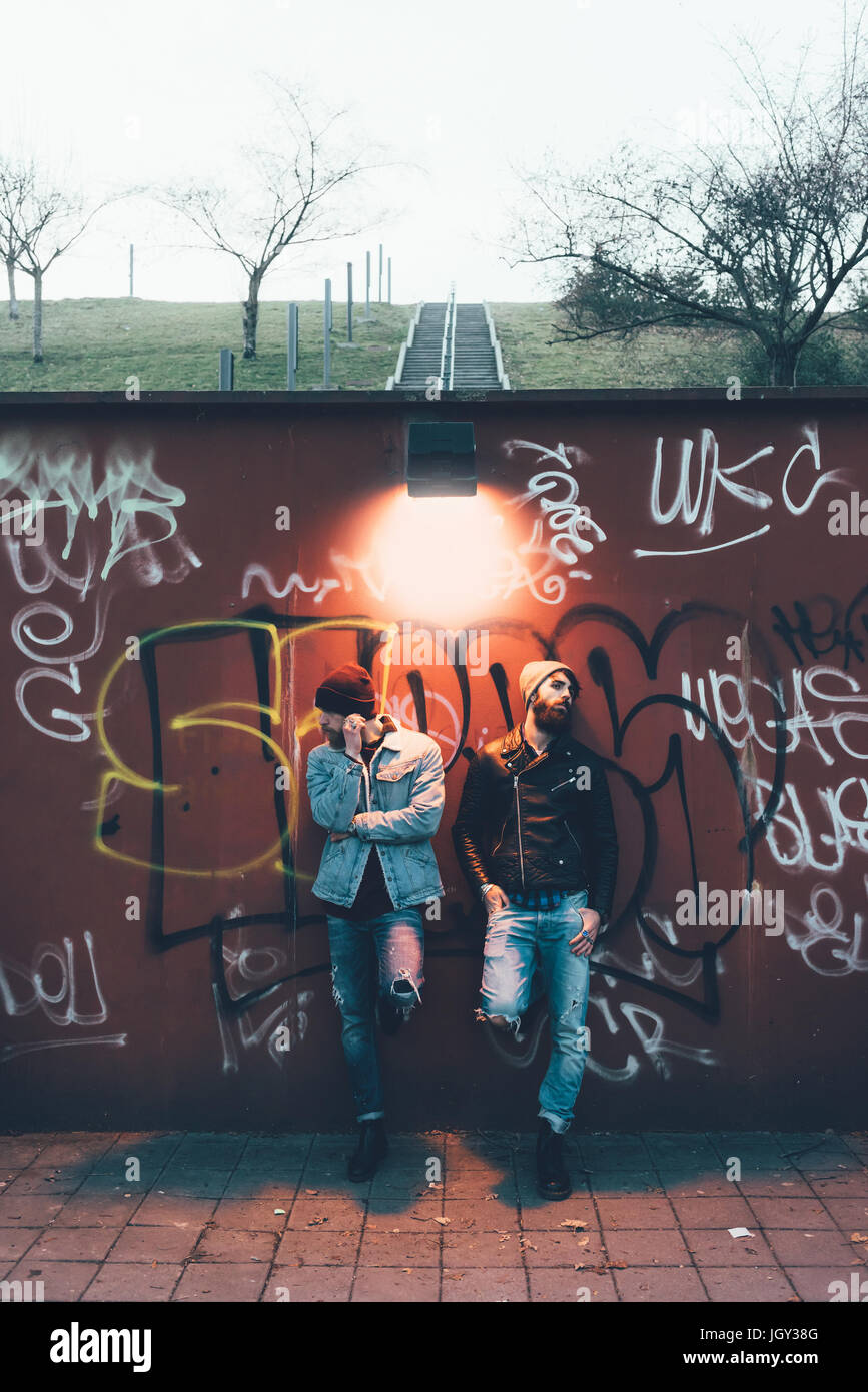Portrait of two cool young male hipsters leaning against graffiti wall Stock Photo