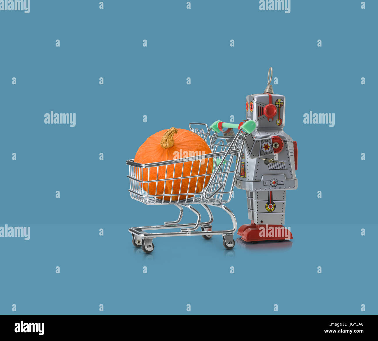 Toy robot pushing miniature shopping trolley with pumpkin against blue background Stock Photo