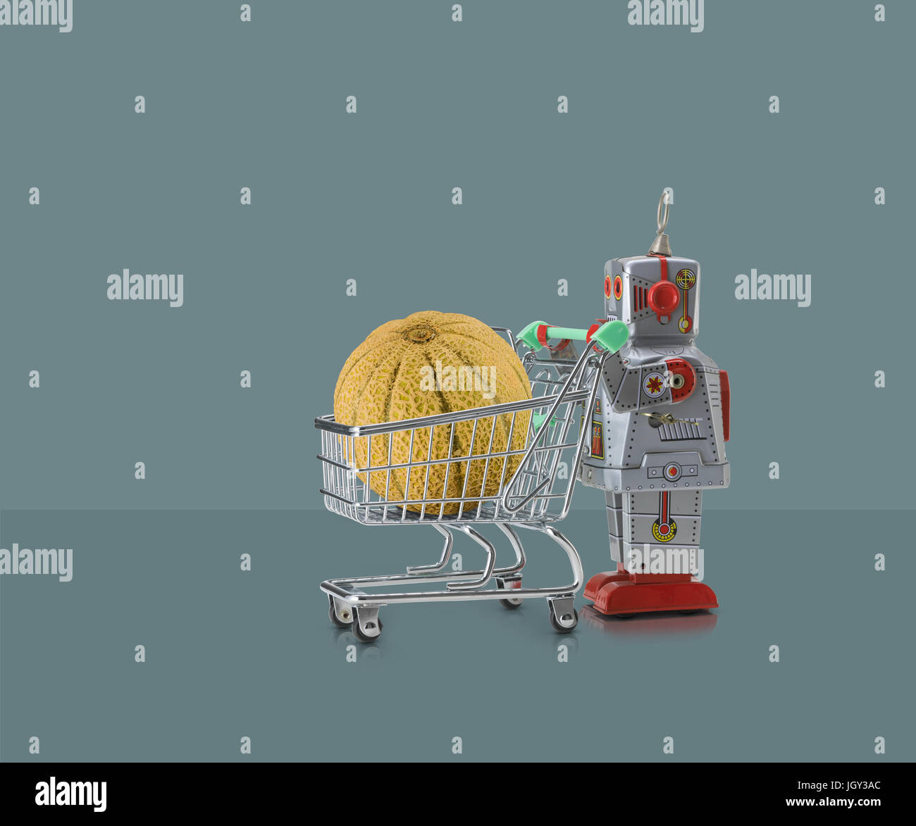 Toy robot pushing miniature shopping trolley with melon against grey background Stock Photo