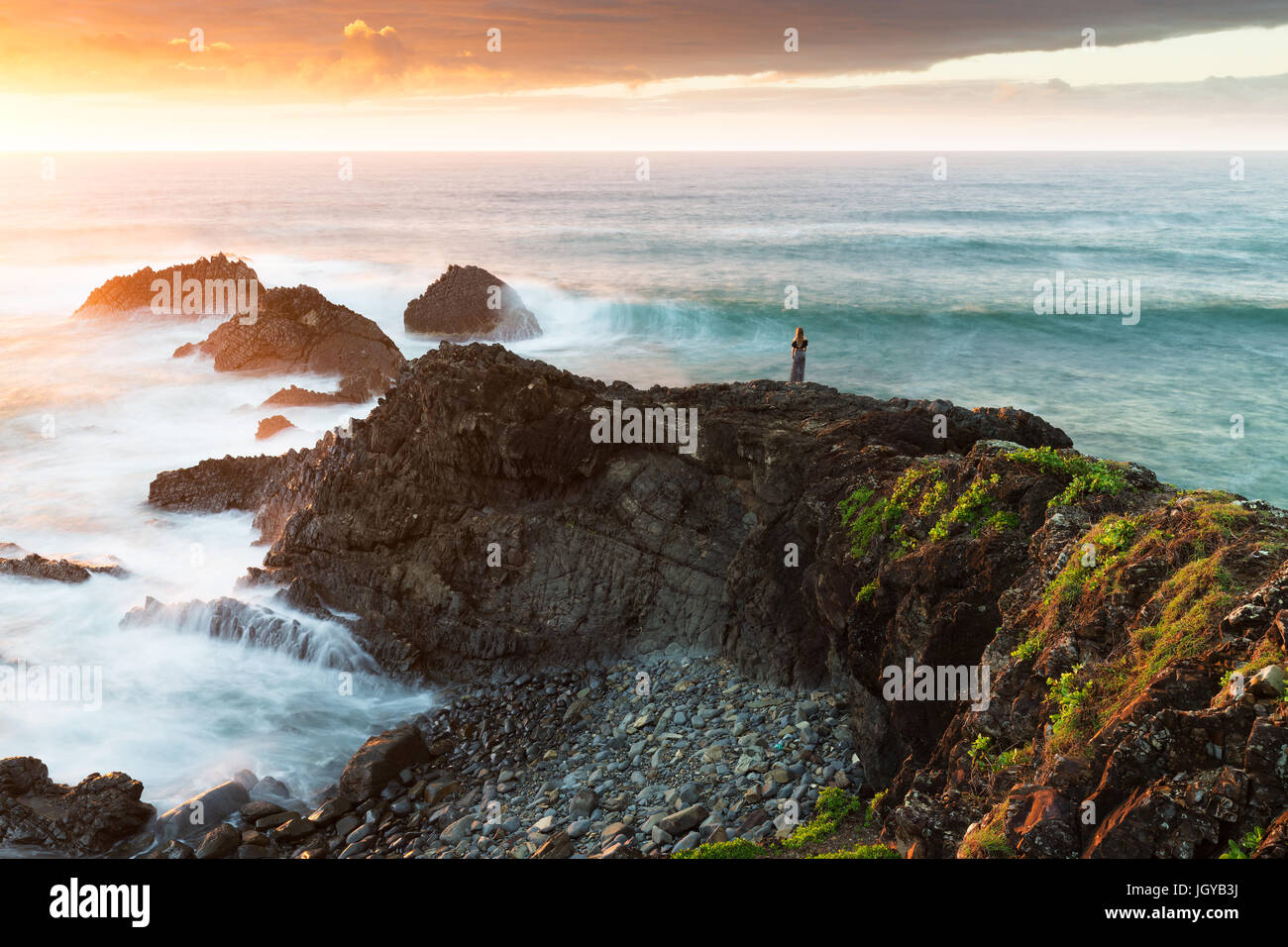 A woman is dwarfed in a majestic coastal scene as the sun rises and highlights the vibrant colours of the landscape and crashing waves. Stock Photo
