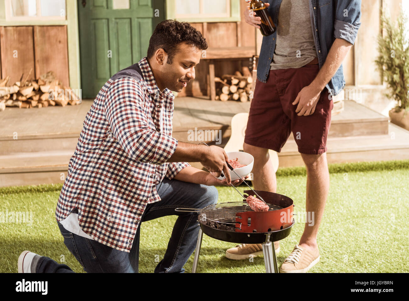 two men grilling burgers while drinking beer together outdoors Stock