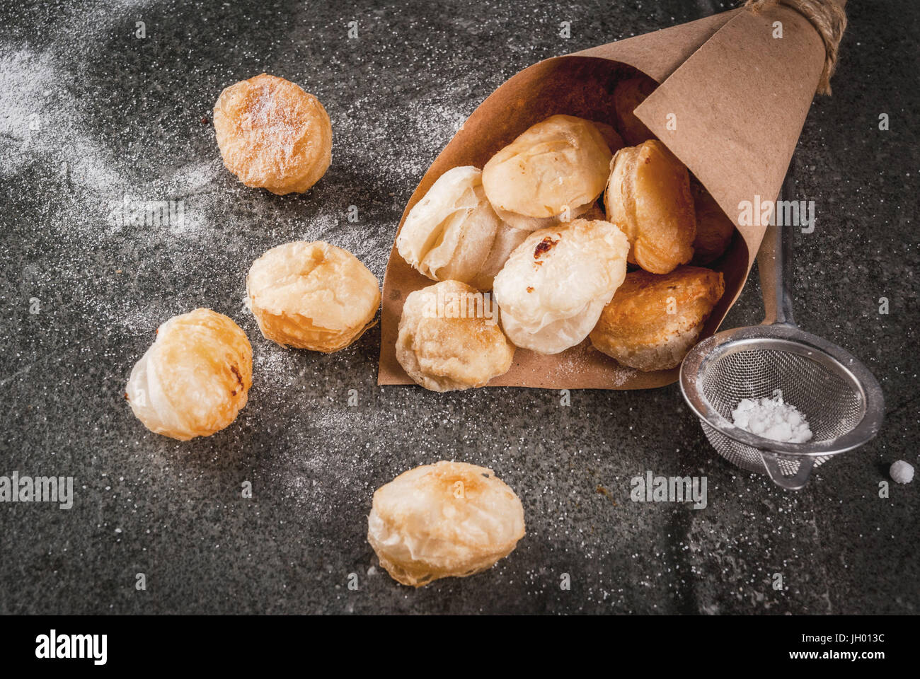 Homemade baking, puff pastries. Trendy food. Cronuts popcorn, puff donuts holes, in paper bag, with powdered sugar. On a dark stone table. Copy space Stock Photo