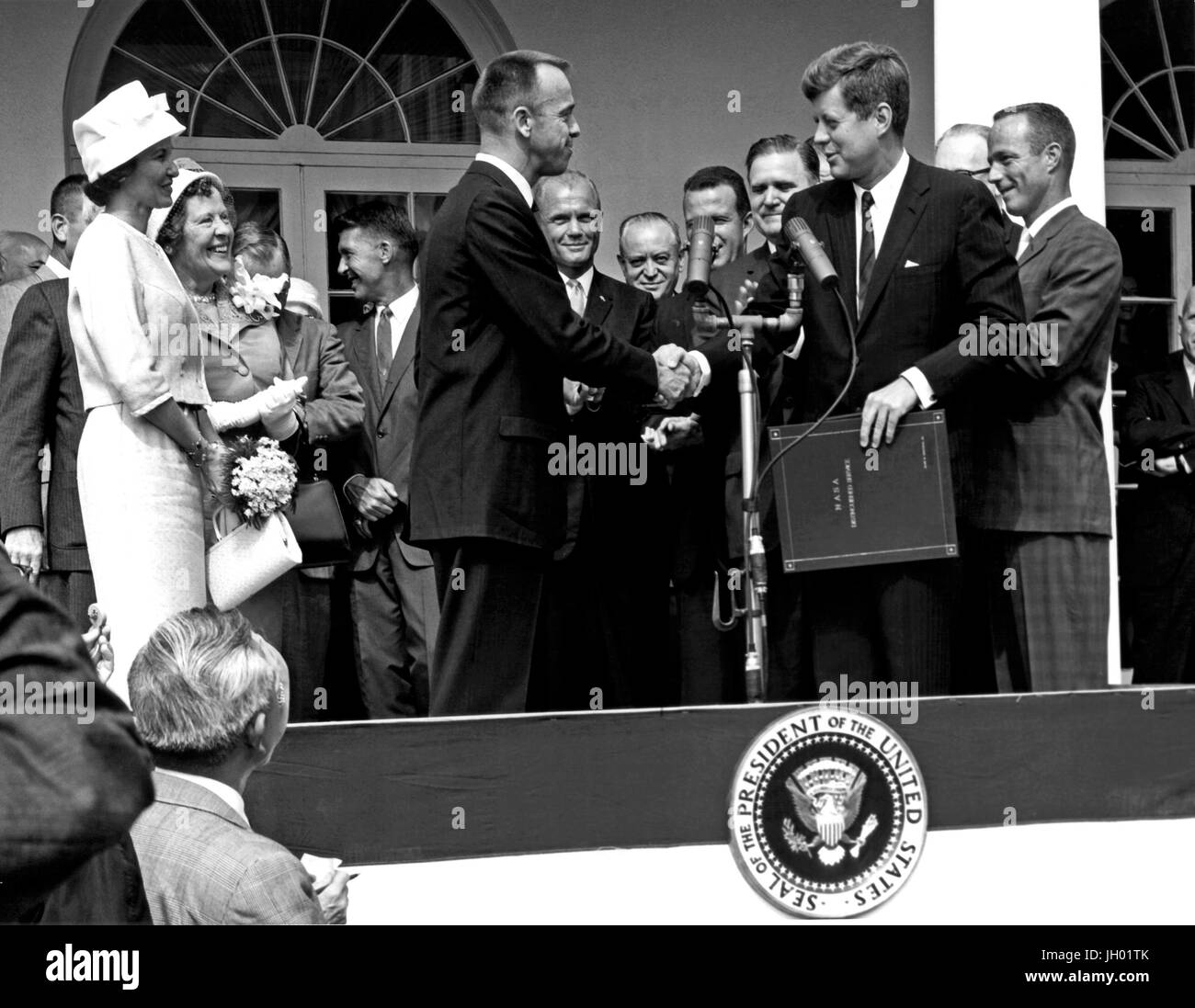 President John F. Kennedy congratulates astronaut Alan B. Shepard, Jr., the first American in space, on his historic May 5th, 1961 ride in the Freedom 7 spacecraft and presents him with the NASA Distinguished Service Award. The ceremony took place on the White House lawn. Shepard's wife, Louise (left in white dress and hat), and his mother were in attendance as well as the other six Mercury astronauts and NASA officals, some visible in the background. Stock Photo