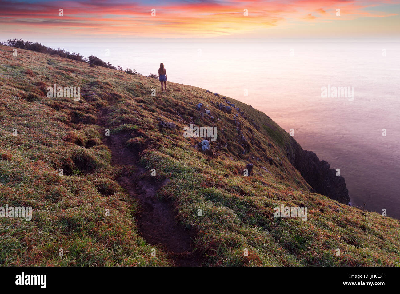 A silhouetted woman walking on a coastal trail, stops to watch a beautiful pink sunrise above the cliff tops near Port Macquarie, Australia. Stock Photo