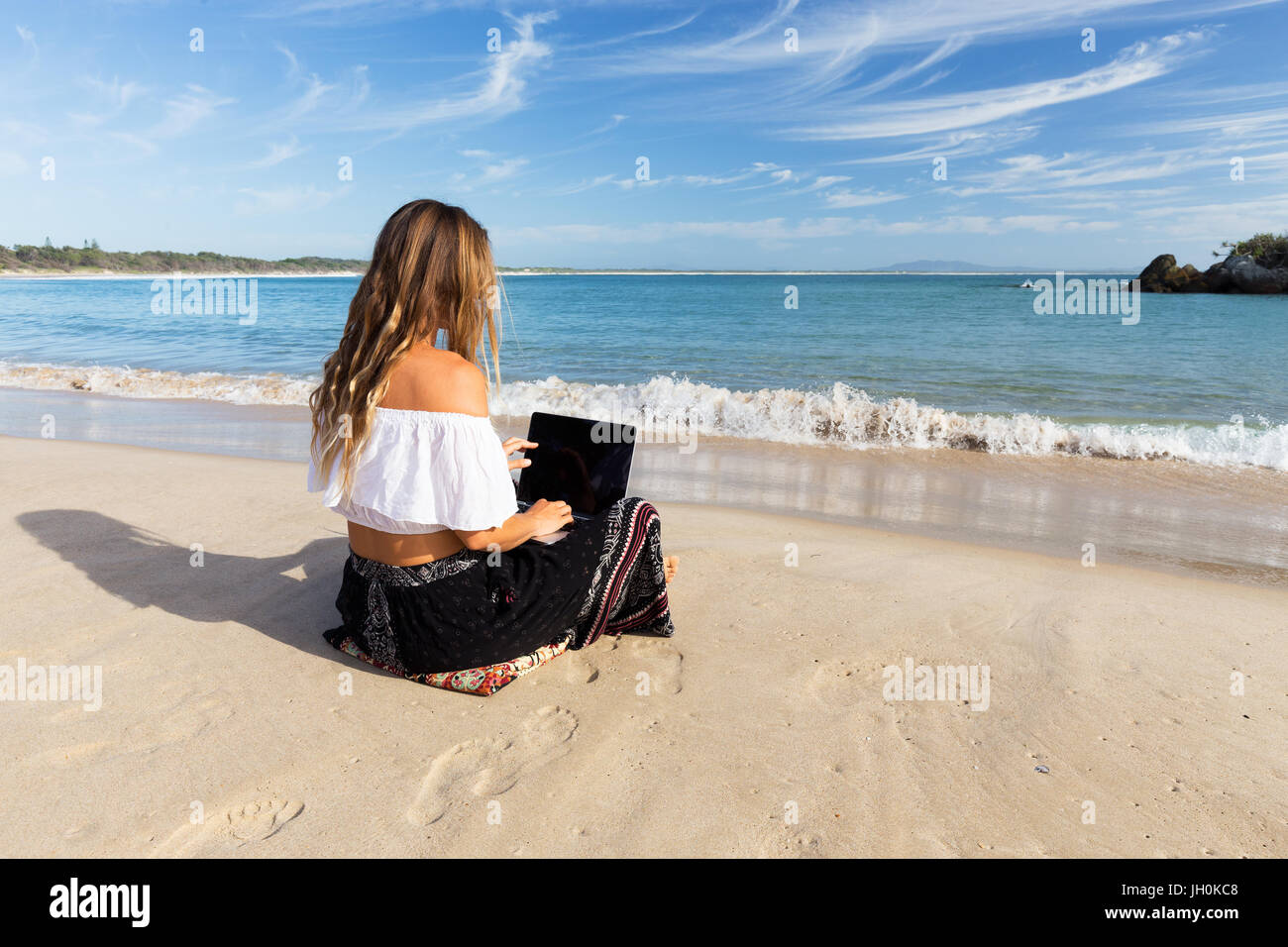 A beautiful young woman sits on the beach beside the sea and crashing waves while working on a laptop. Stock Photo