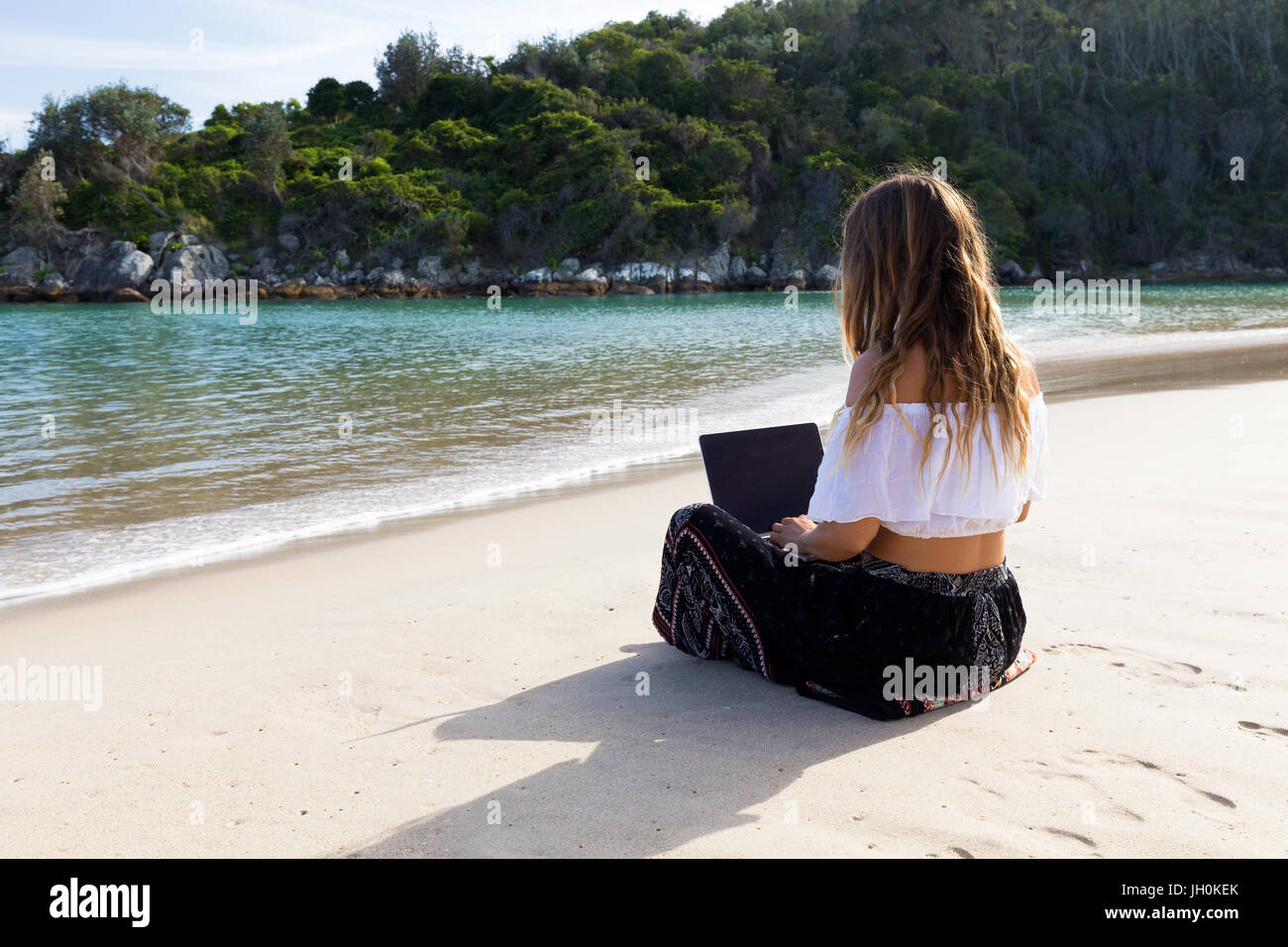 A beautiful young woman sits on the sand beside the crystal clear sea and works on her laptop. Stock Photo