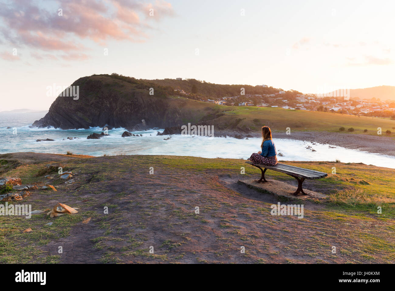 A woman sits on a wooden bench and watches the golden sun light disappearing from the beautiful coastline around her. Stock Photo
