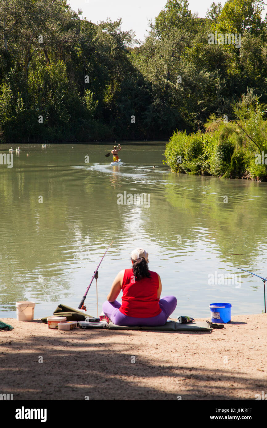 Woman fishing in the river Tagus at Aranjuez in the Madrid province of Spain  watching a paddle boarder go past Stock Photo