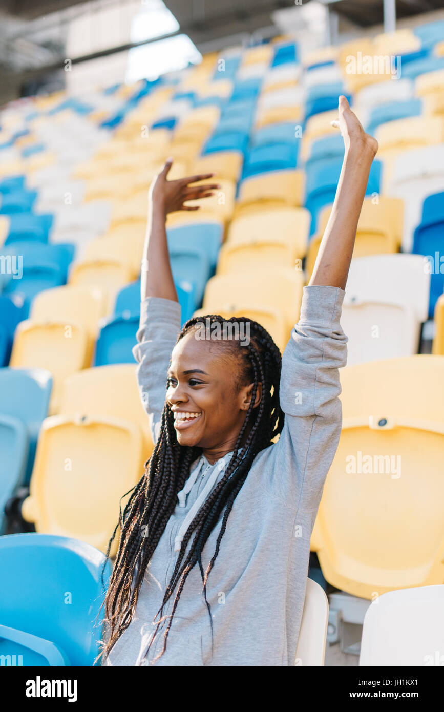 Close-up portrait of the smiling afro-american teenager. The young girl is cheering for the team while sitting on the seats on the stadium. Stock Photo