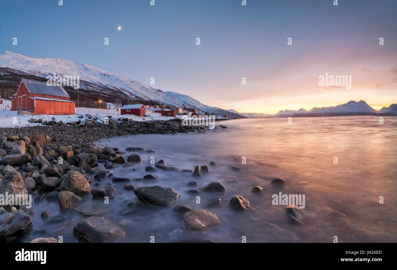 Panorama of typical wood huts called Rorbu framed by fiery sky at sunset and frozen sea, Djupvik, Lyngen Alps, Troms, Norway Stock Photo
