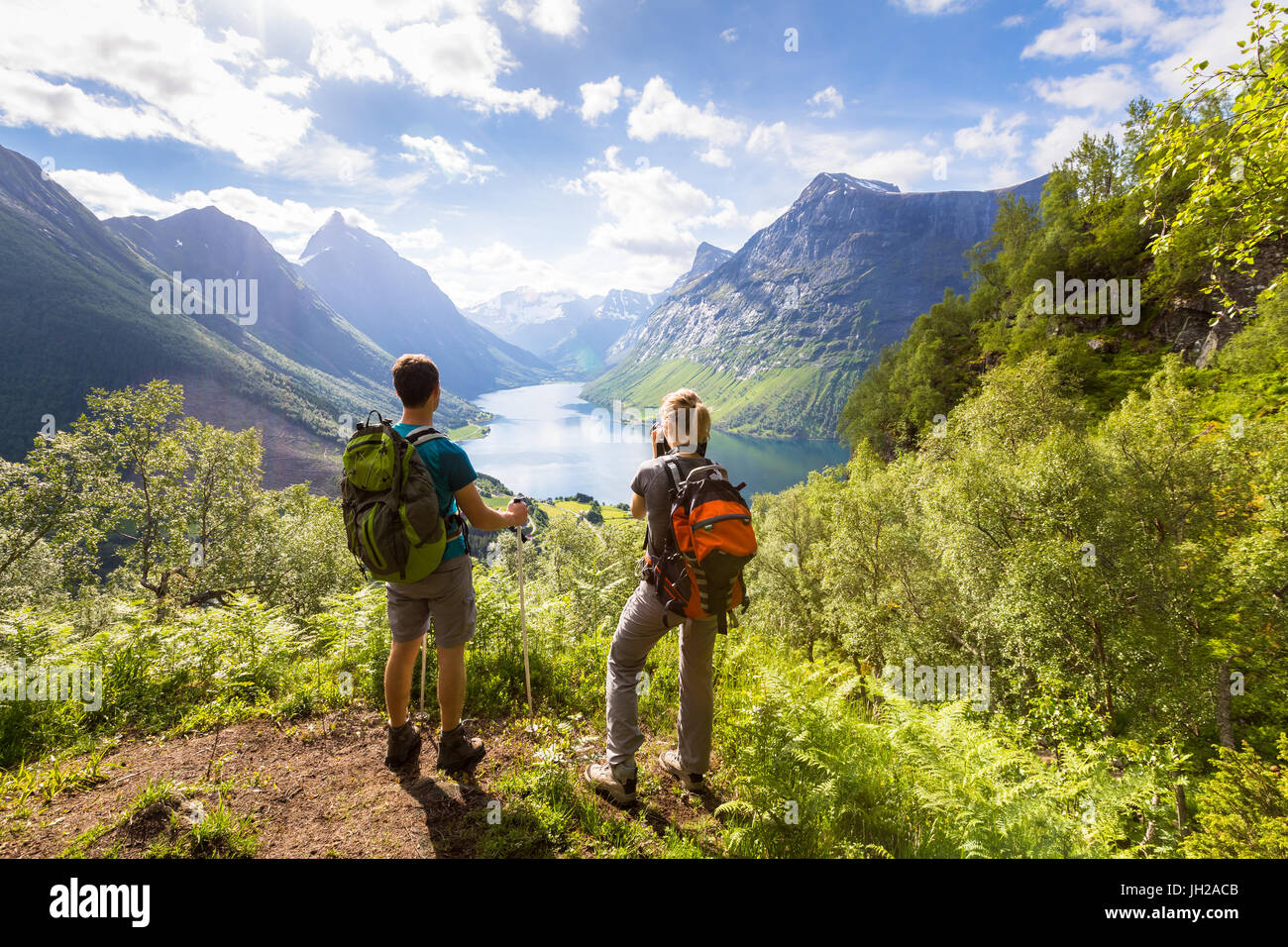 Two hikers at viewpoint in the mountains enjoying beautiful view of the valley with a lake and sunny warm weather in summer, green trees around Stock Photo