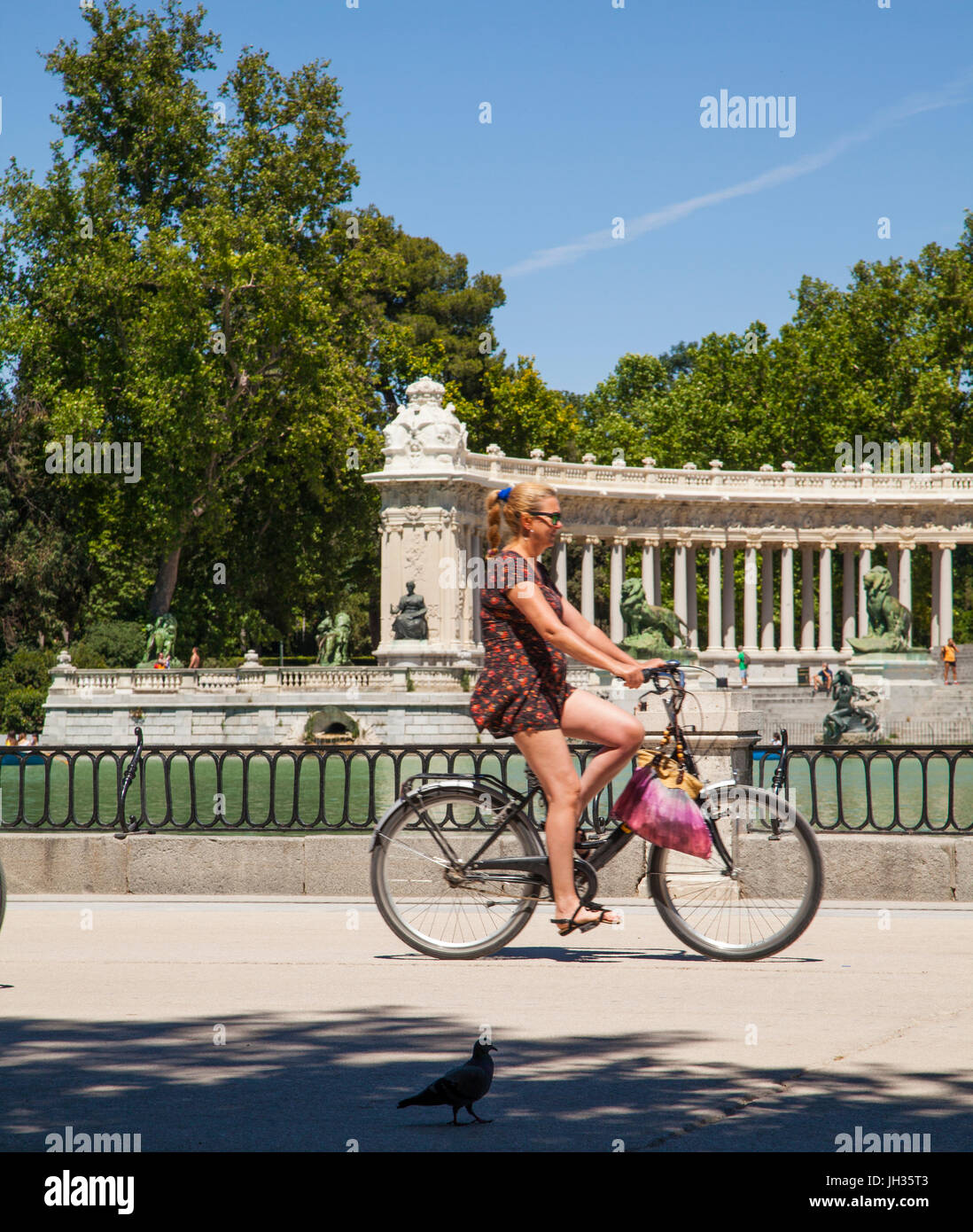 Woman riding a bike in Madrid's Park del Retiro/ Parque del Retiro past the boating lake with the monument to King Alfonso XII n the back ground Stock Photo