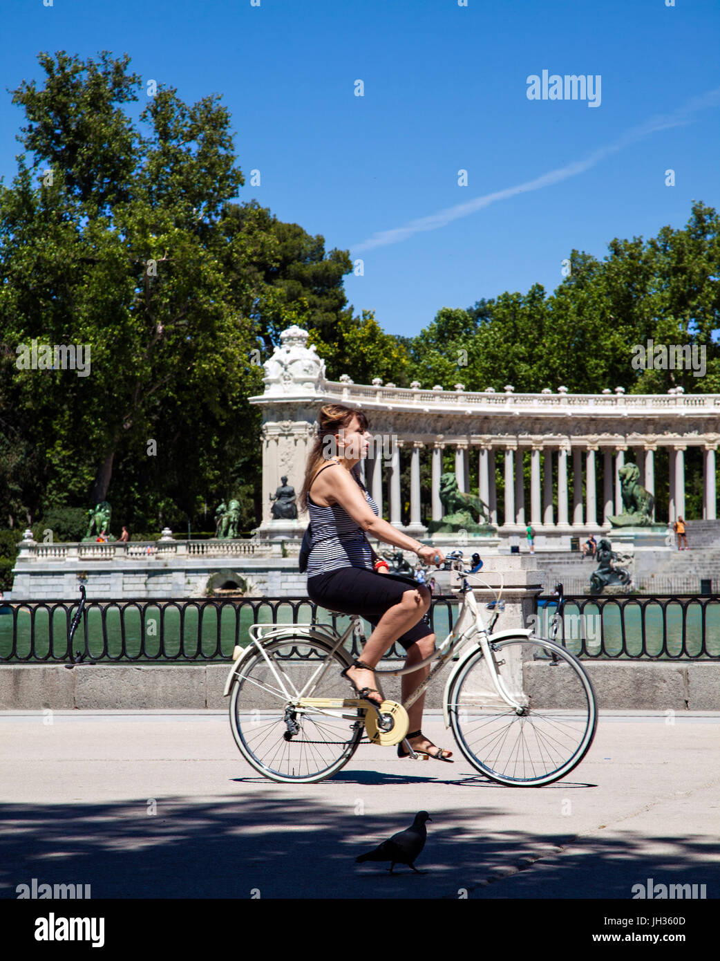 Woman riding a bike in Madrid's Park del Retiro/ Parque del Retiro past the boating lake with the monument to King Alfonso XII in the back ground Stock Photo