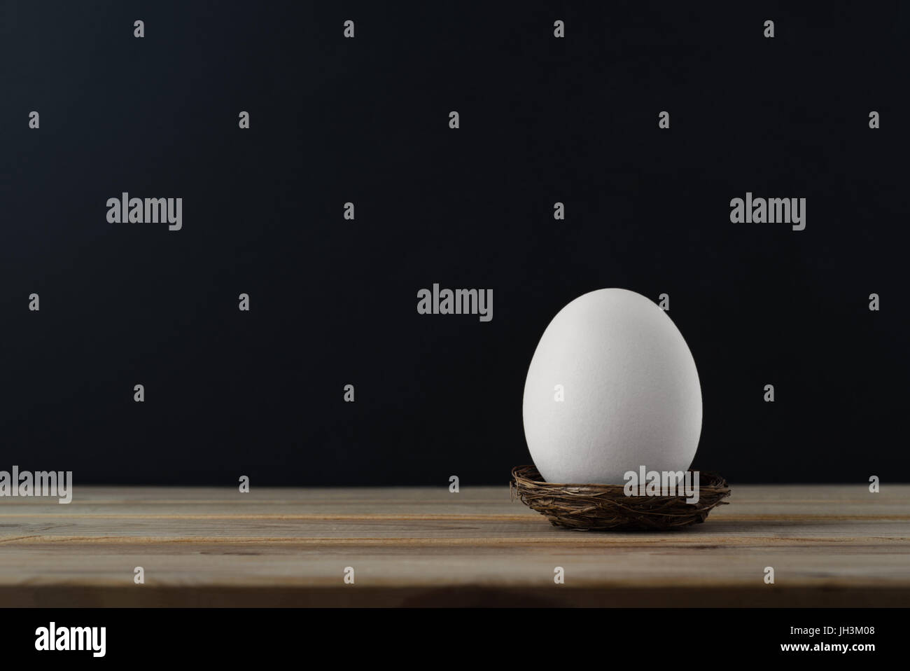 An upright chicken's egg (whitened) in small nest on wood plank table.  Black chalkboard background provides copy space. Stock Photo
