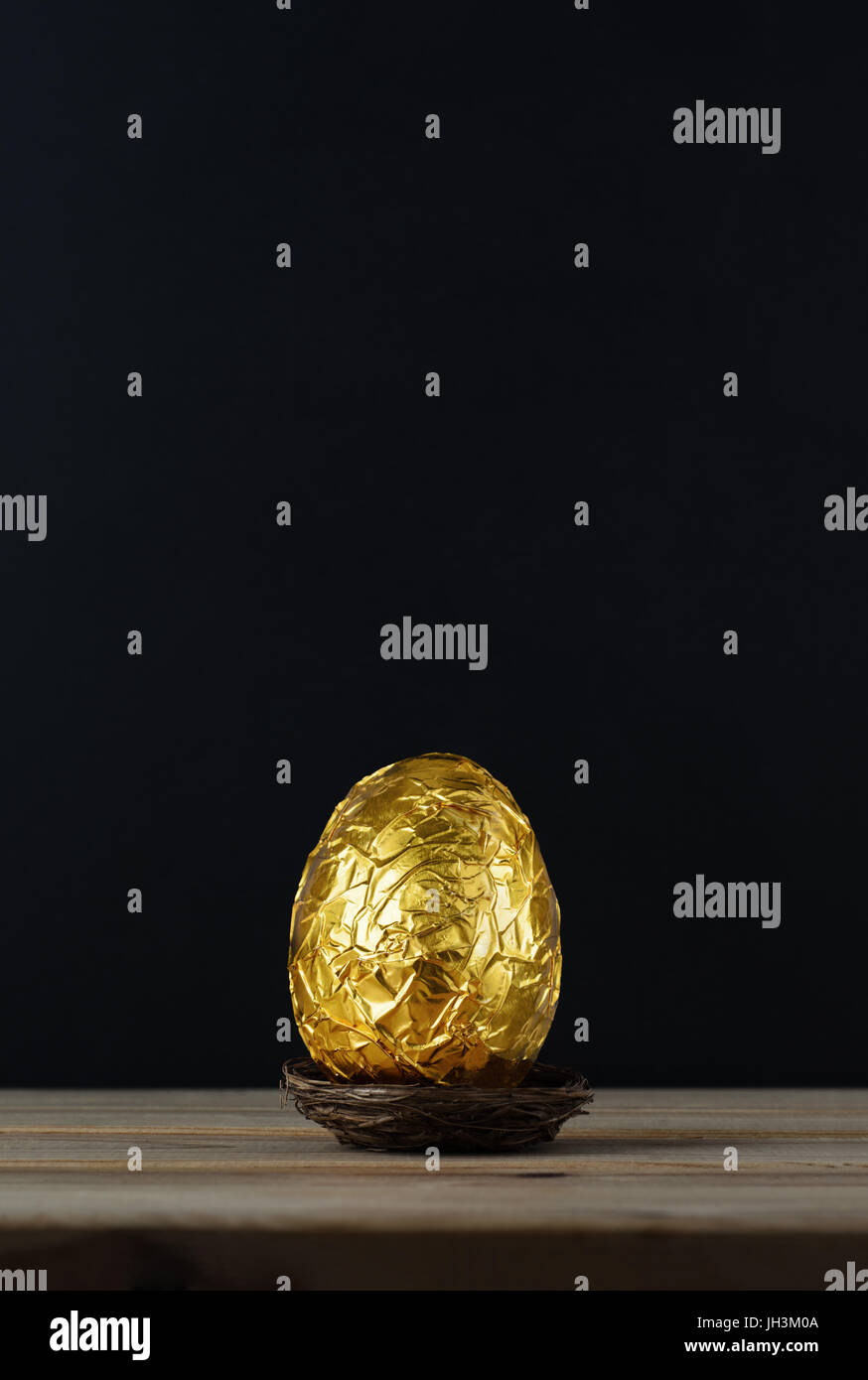 Easter concept.  A chocolate egg, wrapped in crinkled metallic gold foil.  Presented in a small nest on a wood plank table with black chalkboard backg Stock Photo