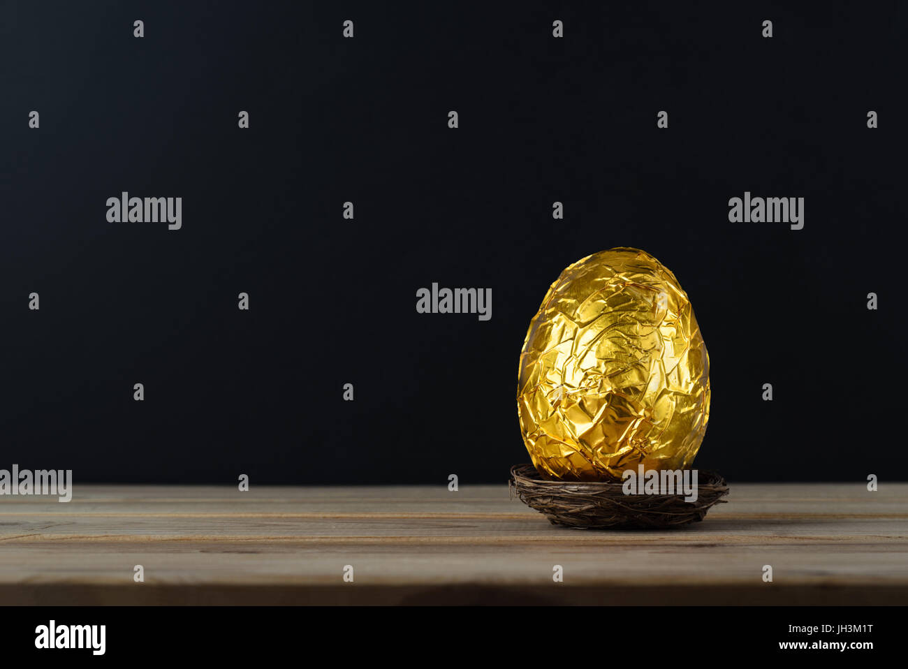 Easter concept.  A chocolate egg, wrapped in crinkled metallic gold foil.  Presented in a small nest on a wood plank table with black chalkboard backg Stock Photo