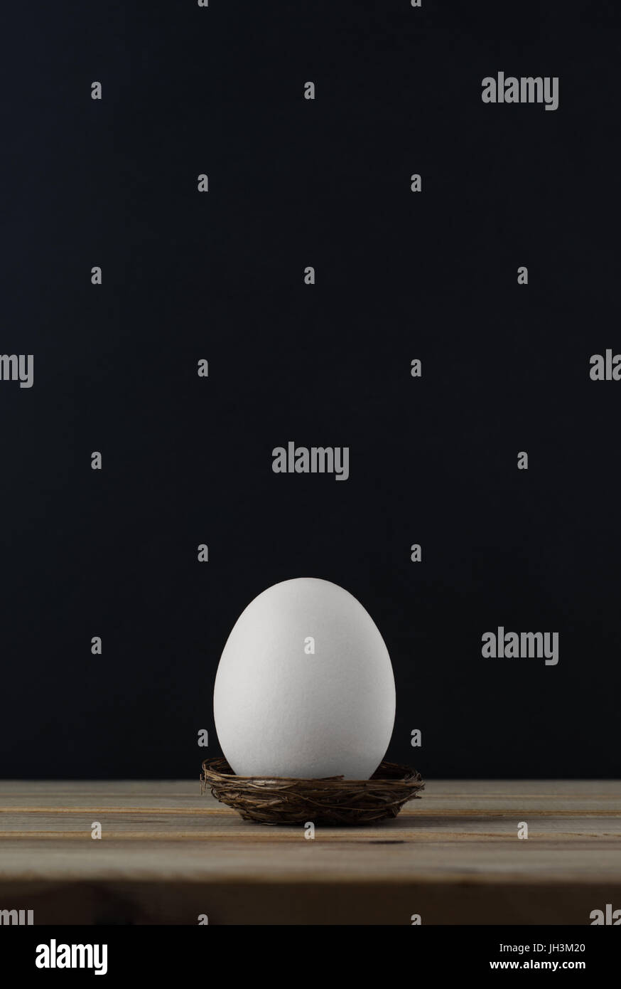 An upright chicken's egg (whitened) in small nest on wood plank table.  Black chalkboard background provides copy space above. Stock Photo