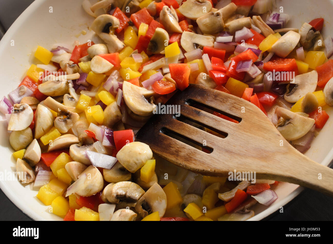 Overhead shot of mushrooms, red onions and red and yellow bell peppers frying in a ceramic pan with old wooden spatula. Stock Photo