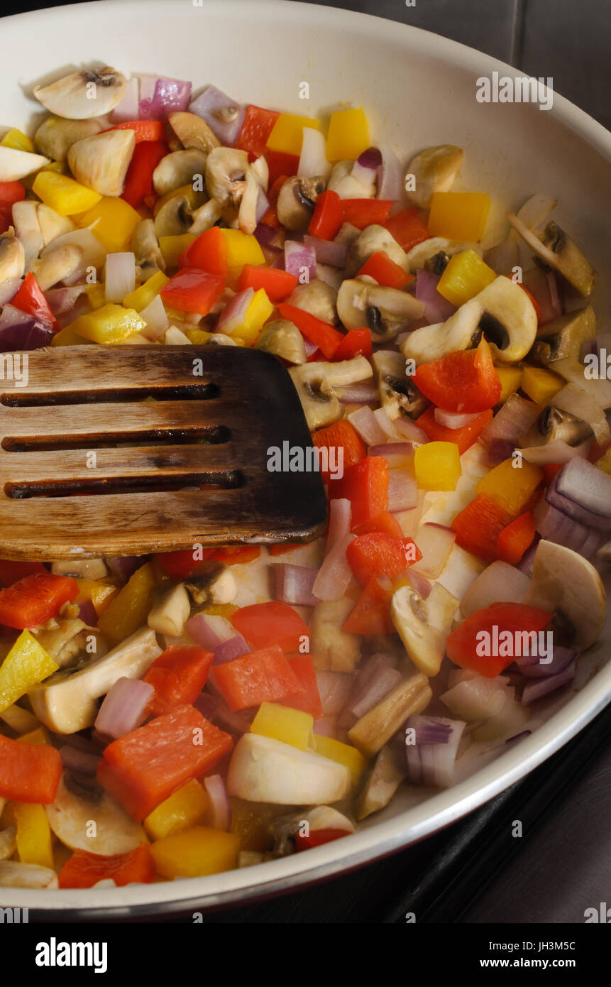 Frying or sauteing proces.  A mixture of fresh vegetables in a ceramic pan on cooker hob with wooden spatula.  Rising steam visible at front of pan. Stock Photo