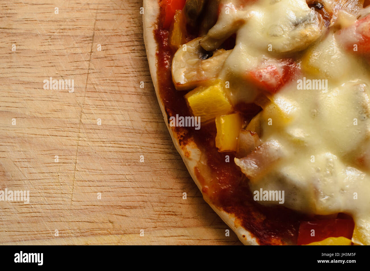 Overhead close up on scratched wooden chopping board of  a cooked vegetarian pizza, with red and yellow peppers, onions, mushrooms and melted mozzarel Stock Photo