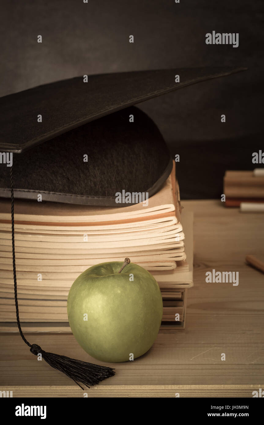 Education Concept.  A wooden teacher's desk with stack of exercise books topped with mortarboard. Apple in the foreground.  Blackboard, duster and cha Stock Photo