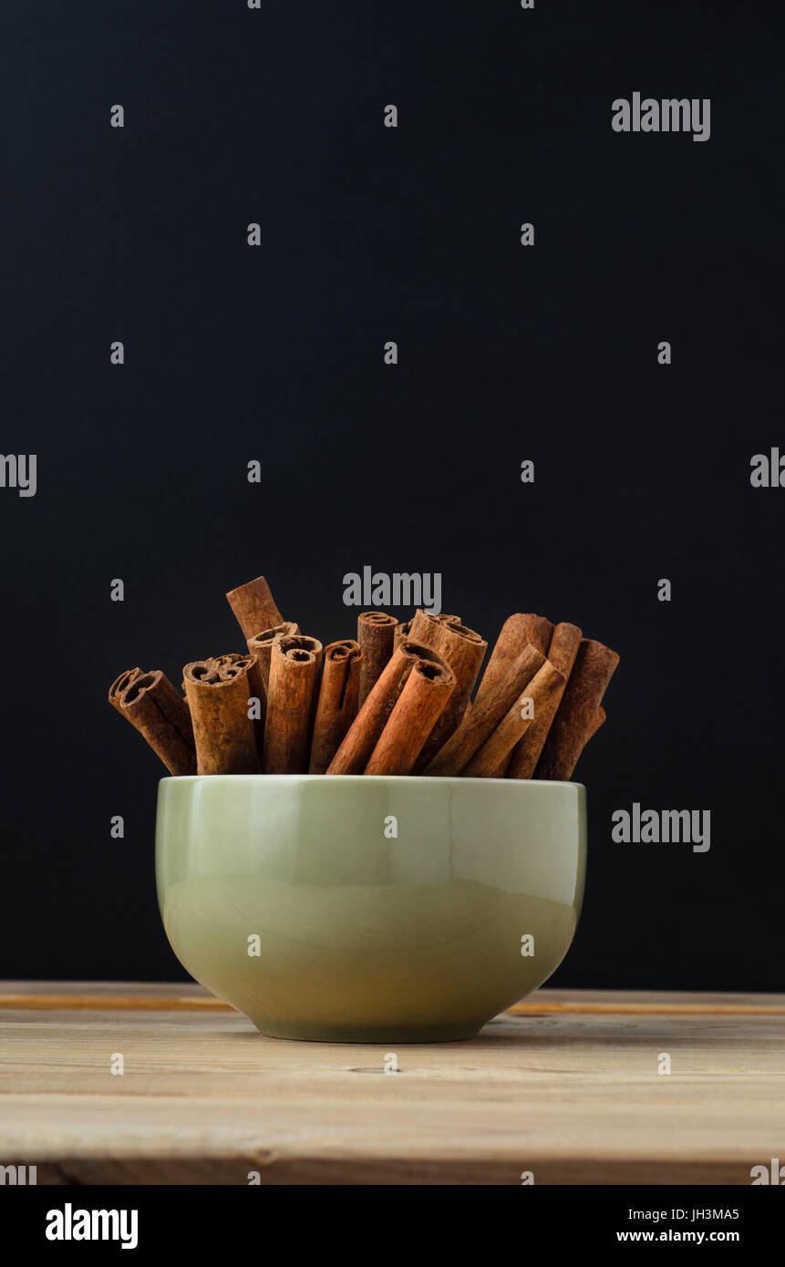 Cinnamon sticks, grouped in a ceramic green bowl on wood planked table, with black chalkboard background providing copy space. Stock Photo