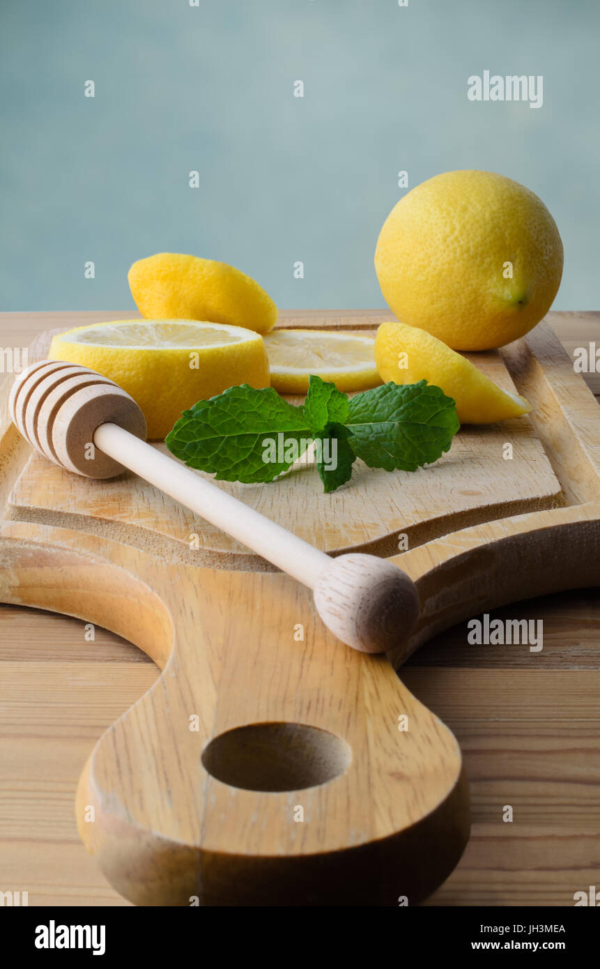 Whole and sliced lemons with mint leaves and honey drizzler on  wooden chopping board with pine kitchen table below. Stock Photo