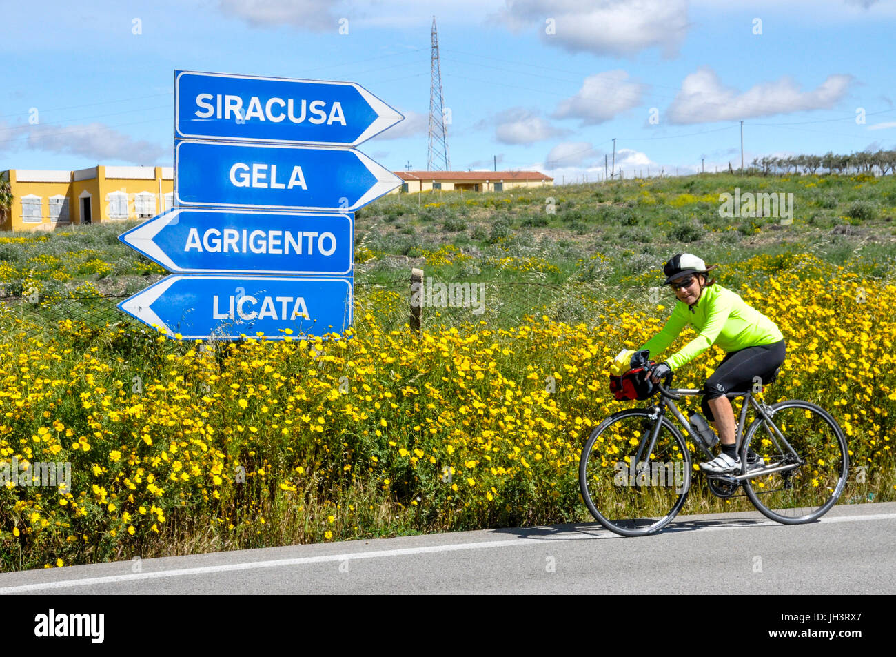 A female touring cyclist  riding on a country road passing a signpost in Sicily, Italy. Stock Photo
