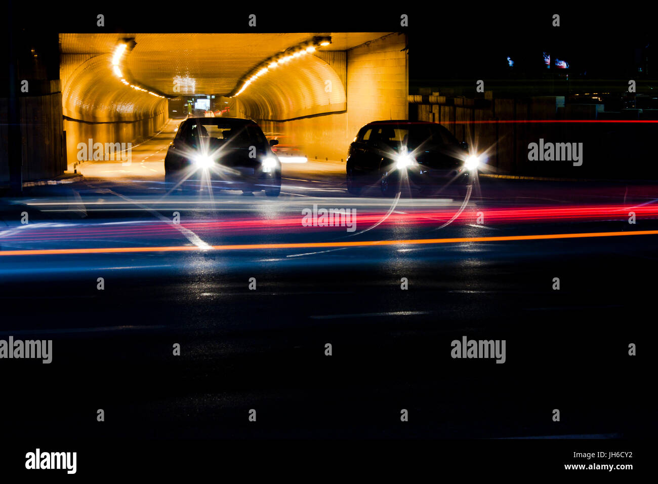 Night traffic on city streets. Cars queued at tunnel exit waiting at intersection while driving vehicles moving past leaving color light trails Stock Photo